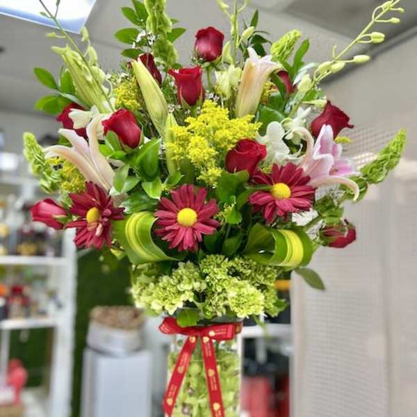 Tall mixed arrangement of red roses, pink lilies, and daisies in a clear cylinder vase with a red ribbon