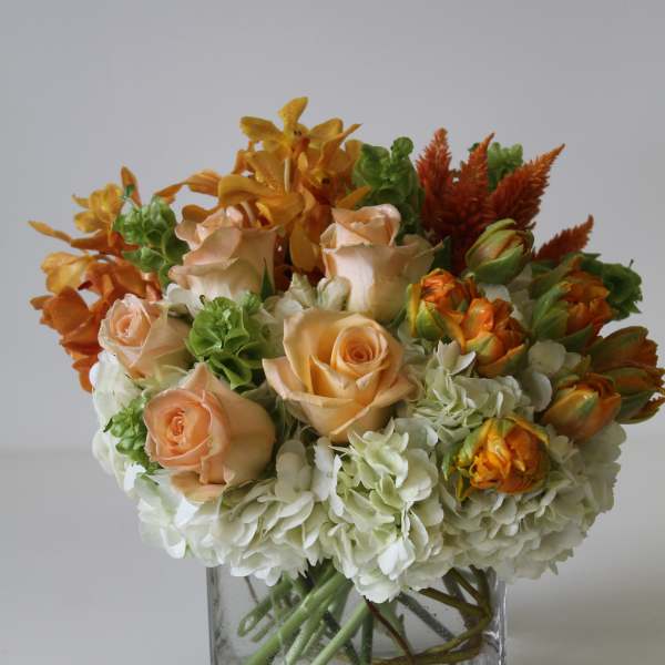 Short arrangement of peach roses, orange blooms, and white hydrangeas in a clear glass cube vase