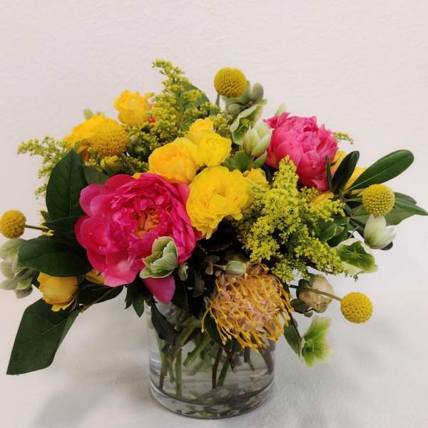 Low arrangement of bright pink peonies and yellow flowers in a clear glass vase