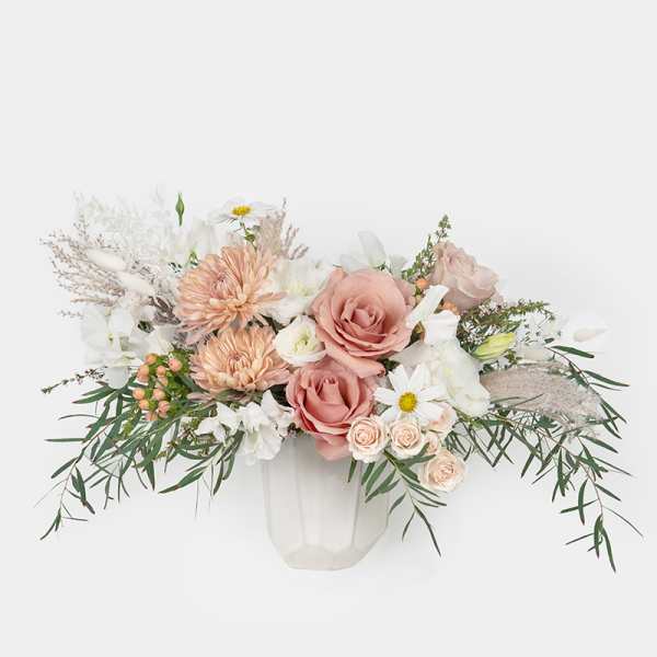 Low arrangement of blush roses, peach mums, and white flowers in a white ceramic vase