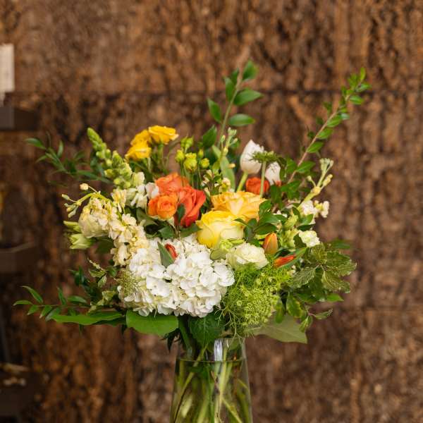 Tall bouquet of yellow and orange roses, white hydrangeas, and mixed flowers in a clear glass vase.
