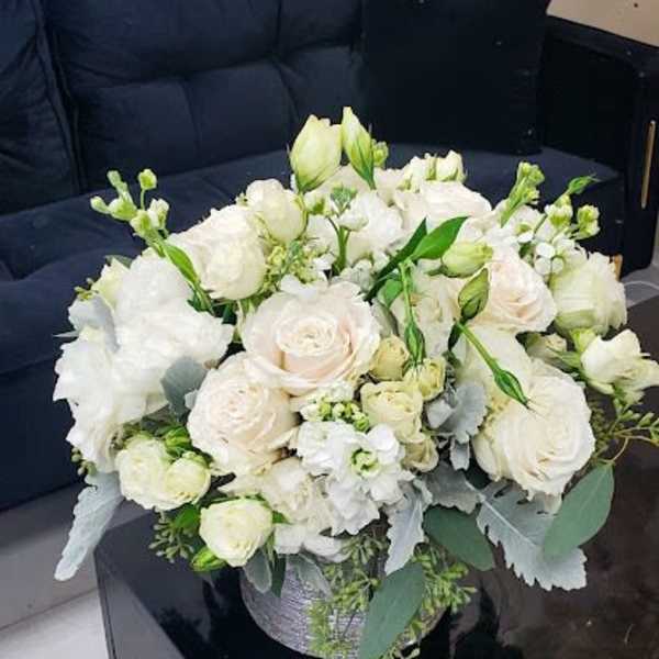 Low round arrangement of white and cream flowers in a silver container on a black table by a dark sofa
