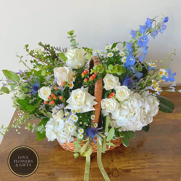 Basket arrangement of white roses, hydrangeas, and blue flowers with berries and a ribbon.