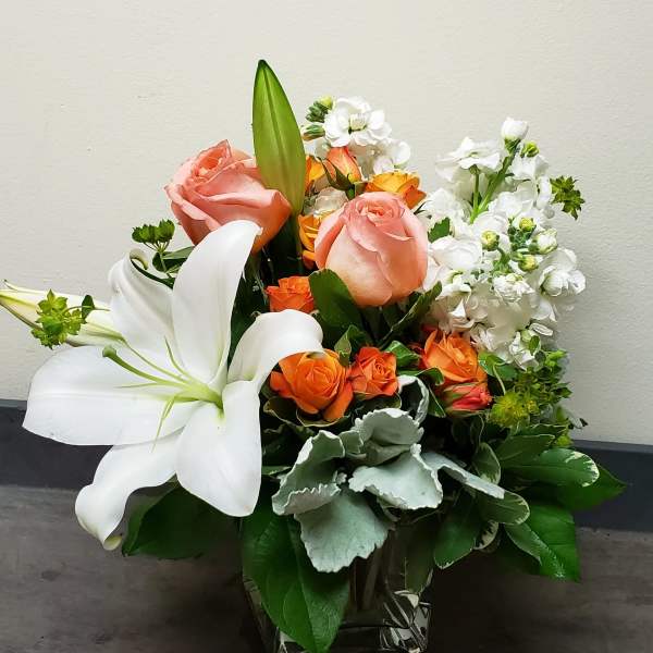 Compact arrangement of white lilies, peach roses, and orange spray roses in a square glass vase