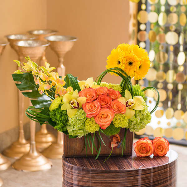 Bright mixed flower arrangement in a wooden box with orange roses and yellow blooms