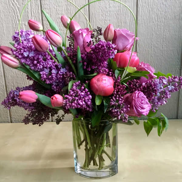 Pink tulips, roses, peonies, and purple lilacs arranged in a clear glass vase