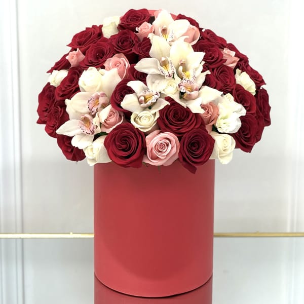 Round arrangement of red, pink, and white roses with orchids in a tall red hatbox-style container