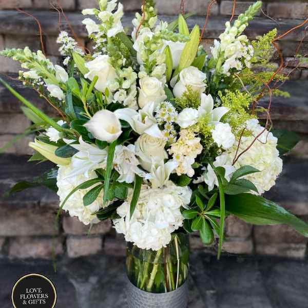 Tall arrangement of white roses, lilies, hydrangeas, and snapdragons in a clear glass vase.