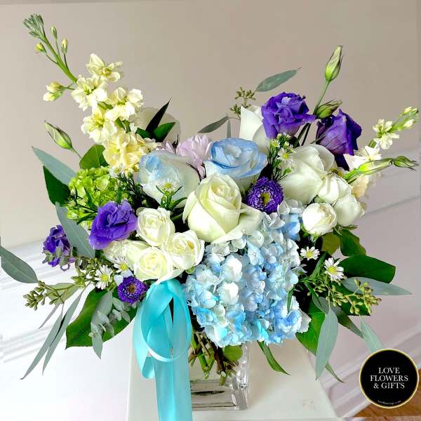 Mixed arrangement of white and blue roses, purple lisianthus, and blue hydrangea in a glass vase with aqua ribbon