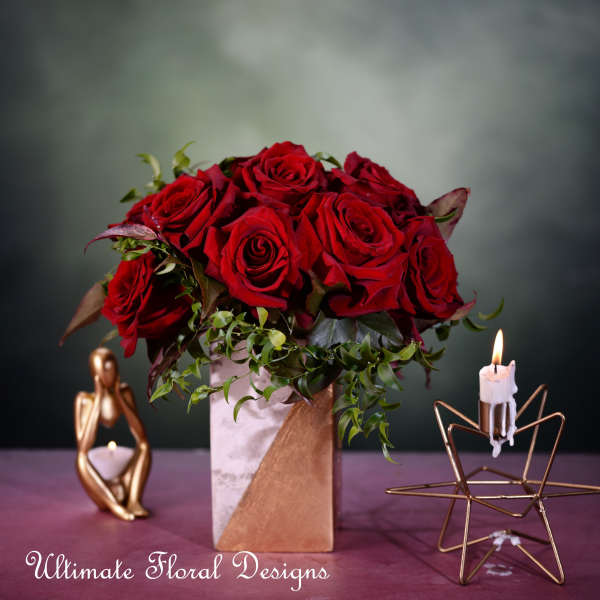 Red roses in a square vase with greenery and candles nearby