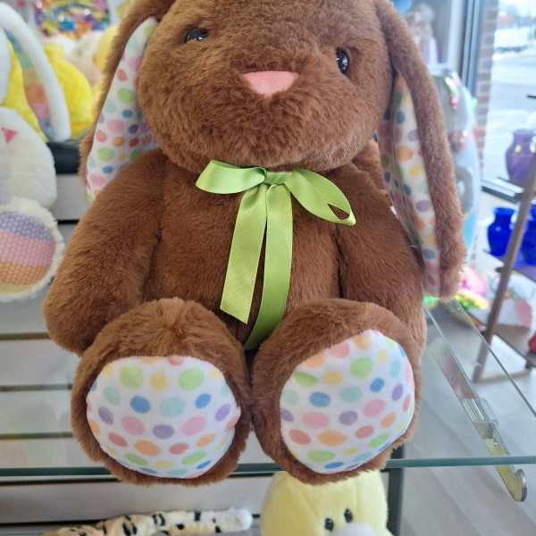 Brown plush bunny with green ribbon and pastel polka dot ears and paws on a glass shelf