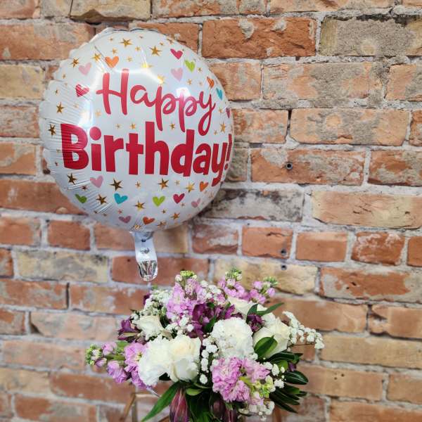 Birthday balloon and small pink and white flower arrangement in a clear vase on a stand.