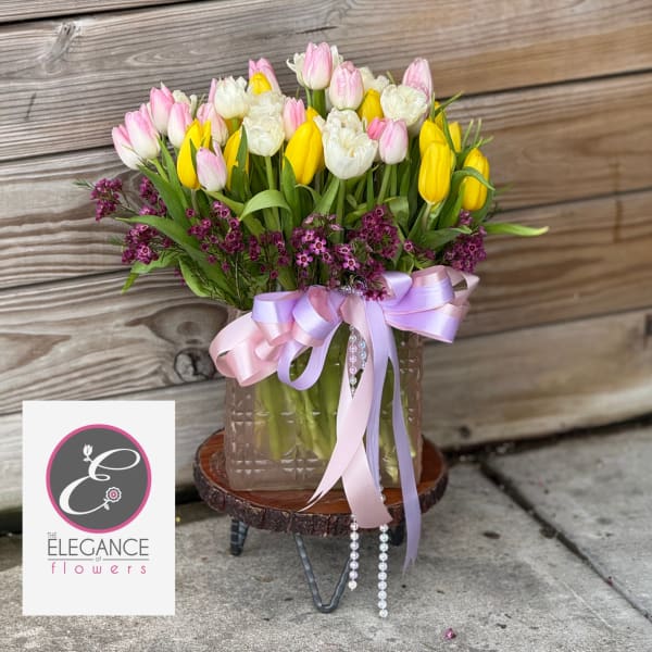 Pastel pink, white, and yellow tulips with purple accents in a glass vase with pink ribbon and pearls.