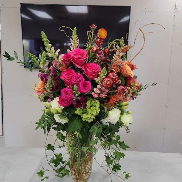 Tall arrangement of hot pink roses and mixed flowers in a clear glass vase with trailing ivy.