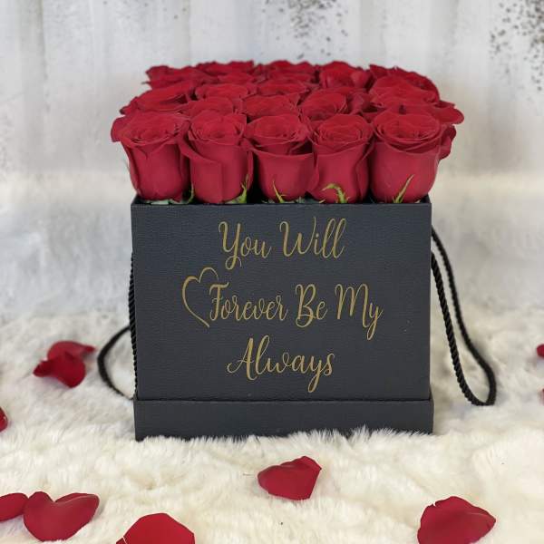 Square black box of red roses with gold script text, surrounded by scattered red rose petals