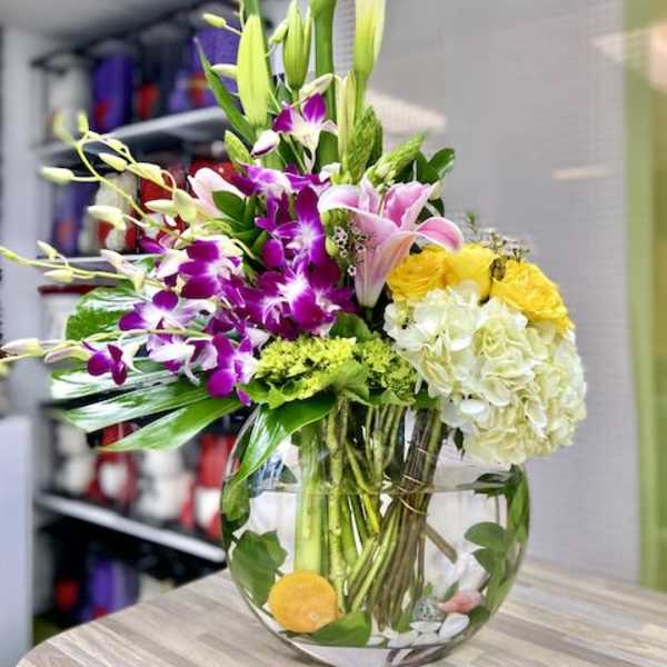 Tall mixed arrangement with white calla lilies, purple orchids, pink lilies, and yellow blooms in a round glass vase