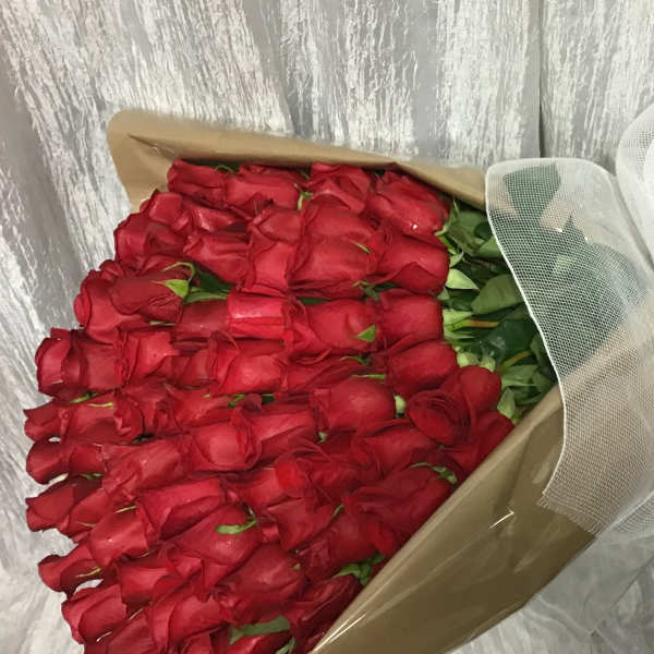 Large bouquet of tightly packed red roses wrapped in kraft paper with white mesh and a red bow.