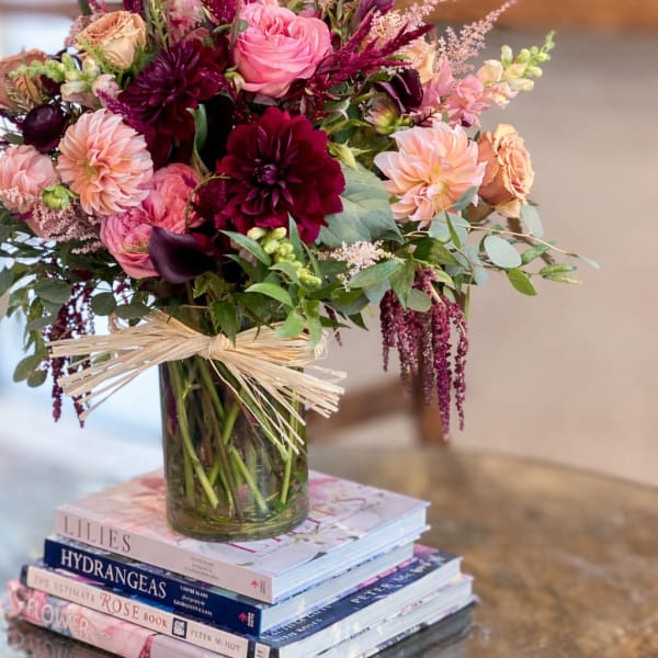Mixed arrangement of pink and burgundy flowers in a glass vase tied with raffia, set on a stack of books.