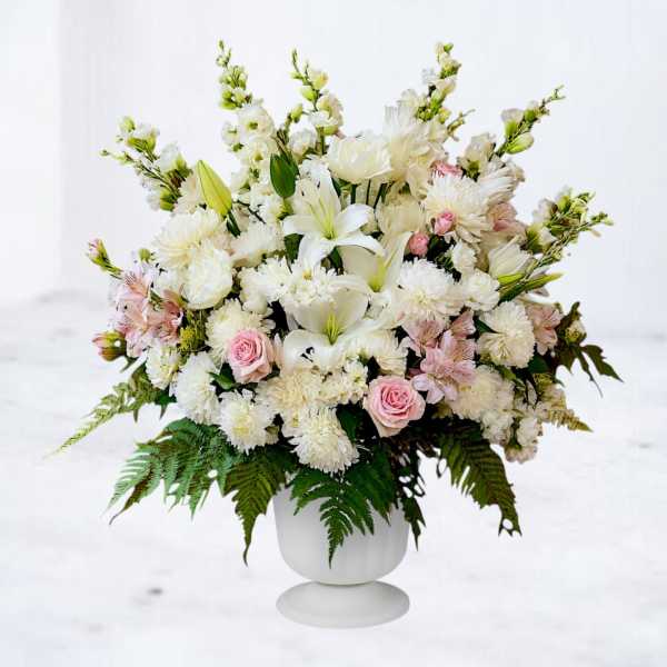 Large white and pink floral arrangement with lilies, roses, and mums in a white pedestal vase