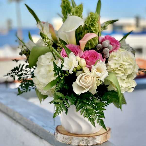 White and pink roses, calla lilies, and hydrangeas arranged in a white vase on a wooden base outdoors