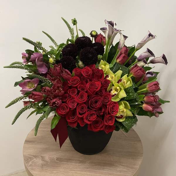Large arrangement of red roses, orchids, tulips, and calla lilies in a black pot on a small round table