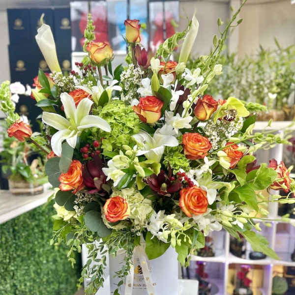 Tall hatbox arrangement with orange roses, white lilies, calla lilies, orchids, and green hydrangea