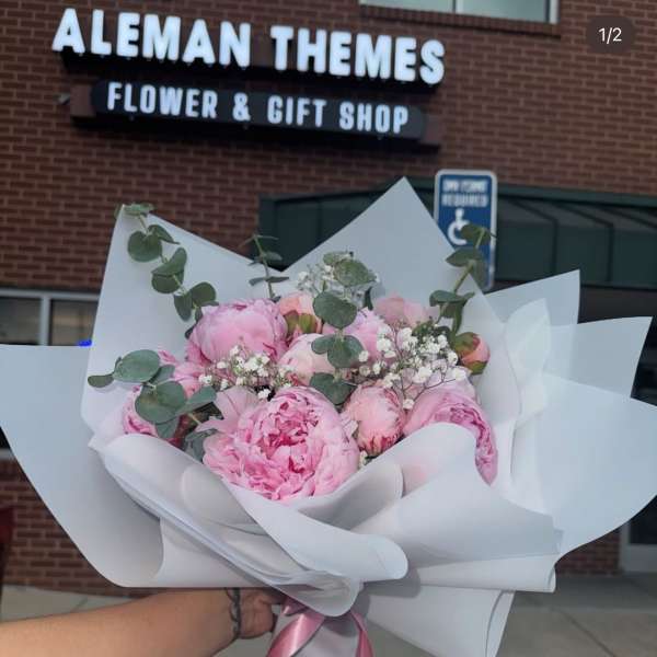 Hand-tied bouquet of pink peonies with white filler flowers wrapped in white paper with a pink ribbon.