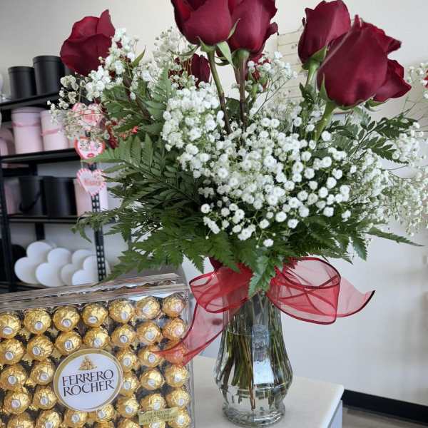 Red roses in a glass vase with baby's breath and a red ribbon, beside a box of Ferrero Rocher chocolates.