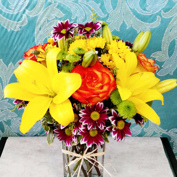 Bright bouquet of yellow lilies, orange roses, and mixed daisies in a clear rectangular vase