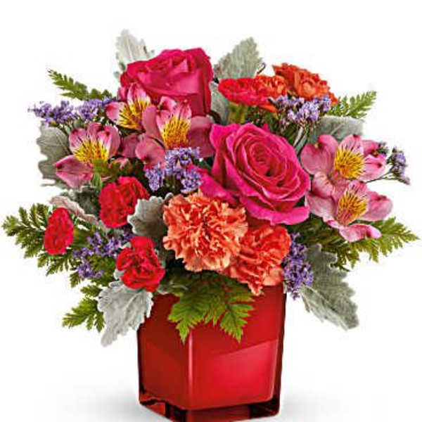 Compact arrangement of pink roses, orange carnations, and mixed blooms in a red cube vase