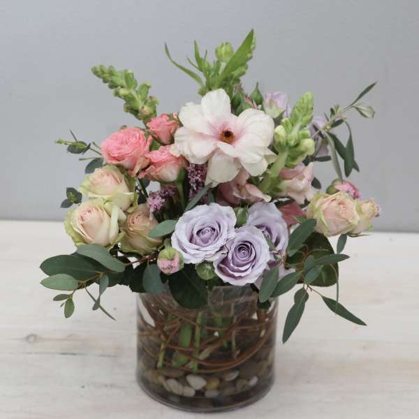 Pastel arrangement of pink and lavender roses, anemone, and snapdragons in a clear glass vase with stones