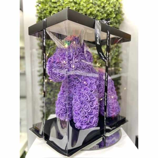 Purple rose-covered teddy bear in a clear display box with a black base and lid.
