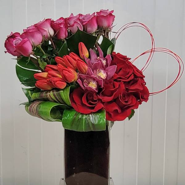 Pink and red floral arrangement in a tall glass vase with heart-shaped wire accents
