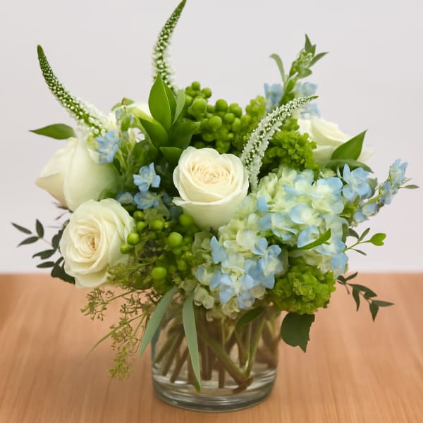 Low round arrangement of white roses, blue hydrangeas, and green accents in a clear glass vase.