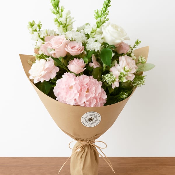 Hand-tied bouquet of soft pink and white flowers wrapped in brown kraft paper.
