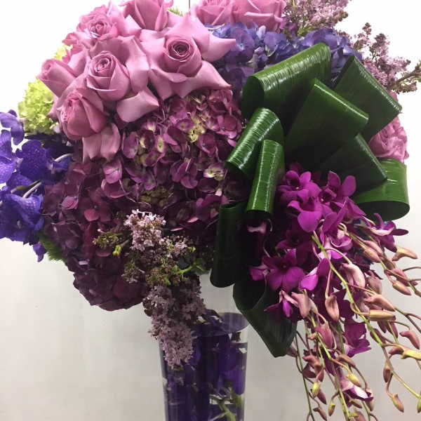 Purple roses and orchids arranged in a clear vase with a large green ribbon