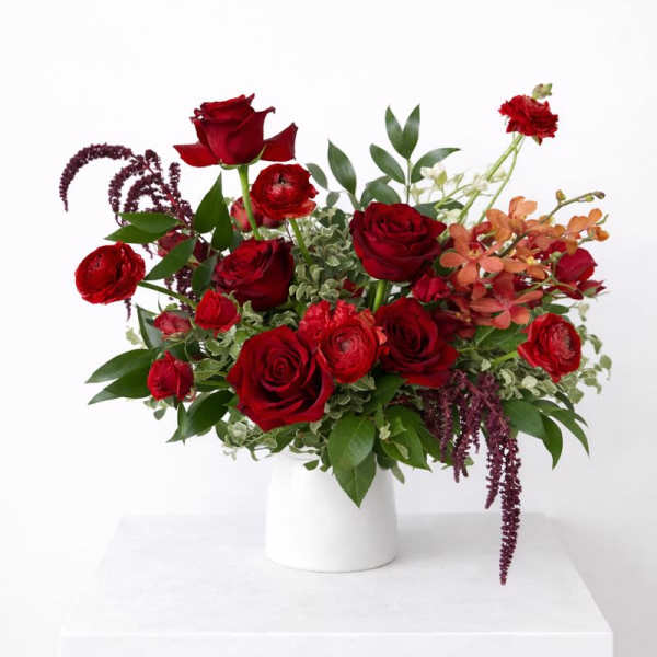 Arrangement of deep red roses, ranunculus, and orange orchids in a white vase