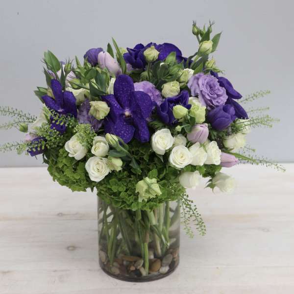 Round arrangement of purple and white flowers with green hydrangea in a clear glass cylinder vase