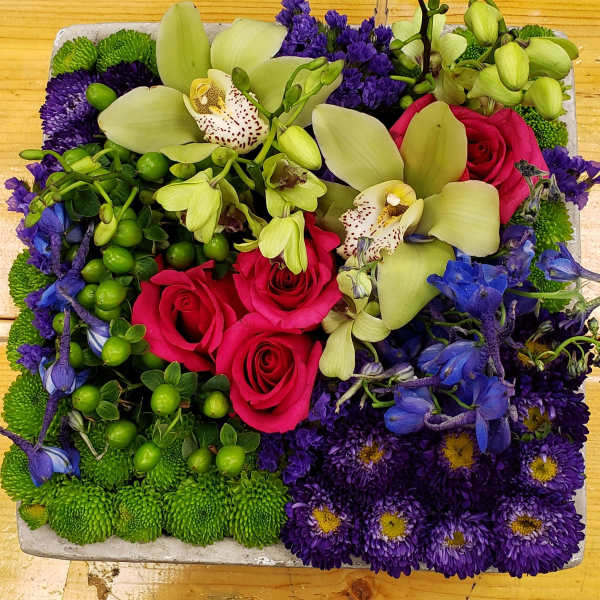 Square arrangement with pink roses, green orchids, purple mums and blue delphinium in a low container