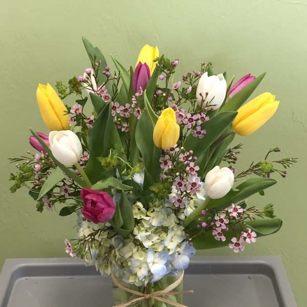 Mixed tulip and hydrangea arrangement with pink filler flowers in a clear glass vase tied with raffia.