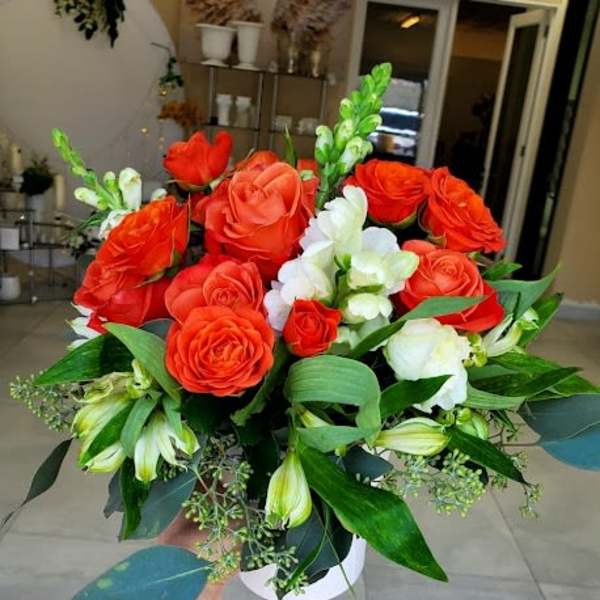 Arrangement of bright orange roses and white flowers with green foliage in a white round vase.