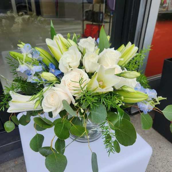 Low arrangement of white roses and lilies with light blue flowers in a clear glass bowl vase