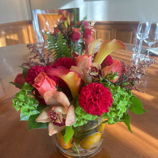 Low glass vase of red dahlias, peach roses, calla lilies, orchids, and green blooms on a wood table