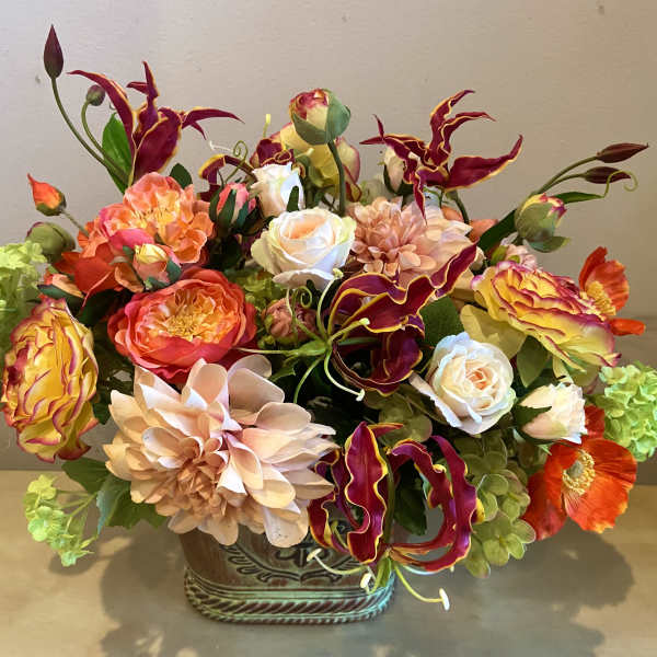 Lush centerpiece of orange, peach, and yellow blooms with burgundy accents in a decorative ceramic container