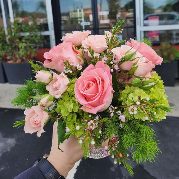 Small bouquet of pink roses with green hydrangea in a clear glass vase held outdoors.