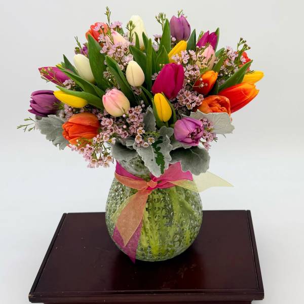 Mixed-color tulip arrangement with pink filler flowers in a green textured vase tied with a ribbon.