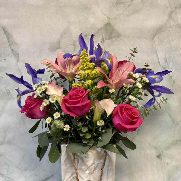Mixed bouquet of pink roses, lilies, and purple irises in a white ceramic bag-style vase