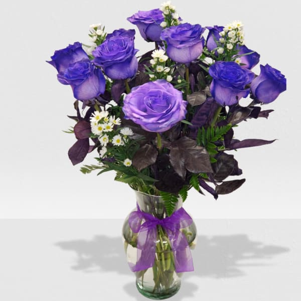 Tall arrangement of purple roses with white daisies and dark purple foliage in a glass vase with a purple bow