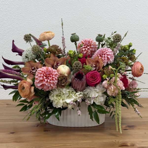 Low oval arrangement of pink dahlias, roses, calla lilies and mixed blooms in a white ceramic vase.