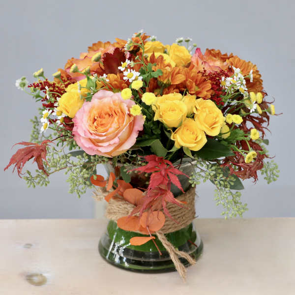 Low glass vase of yellow roses, peach rose, orange mums, and small white daisies with rustic twine wrap.